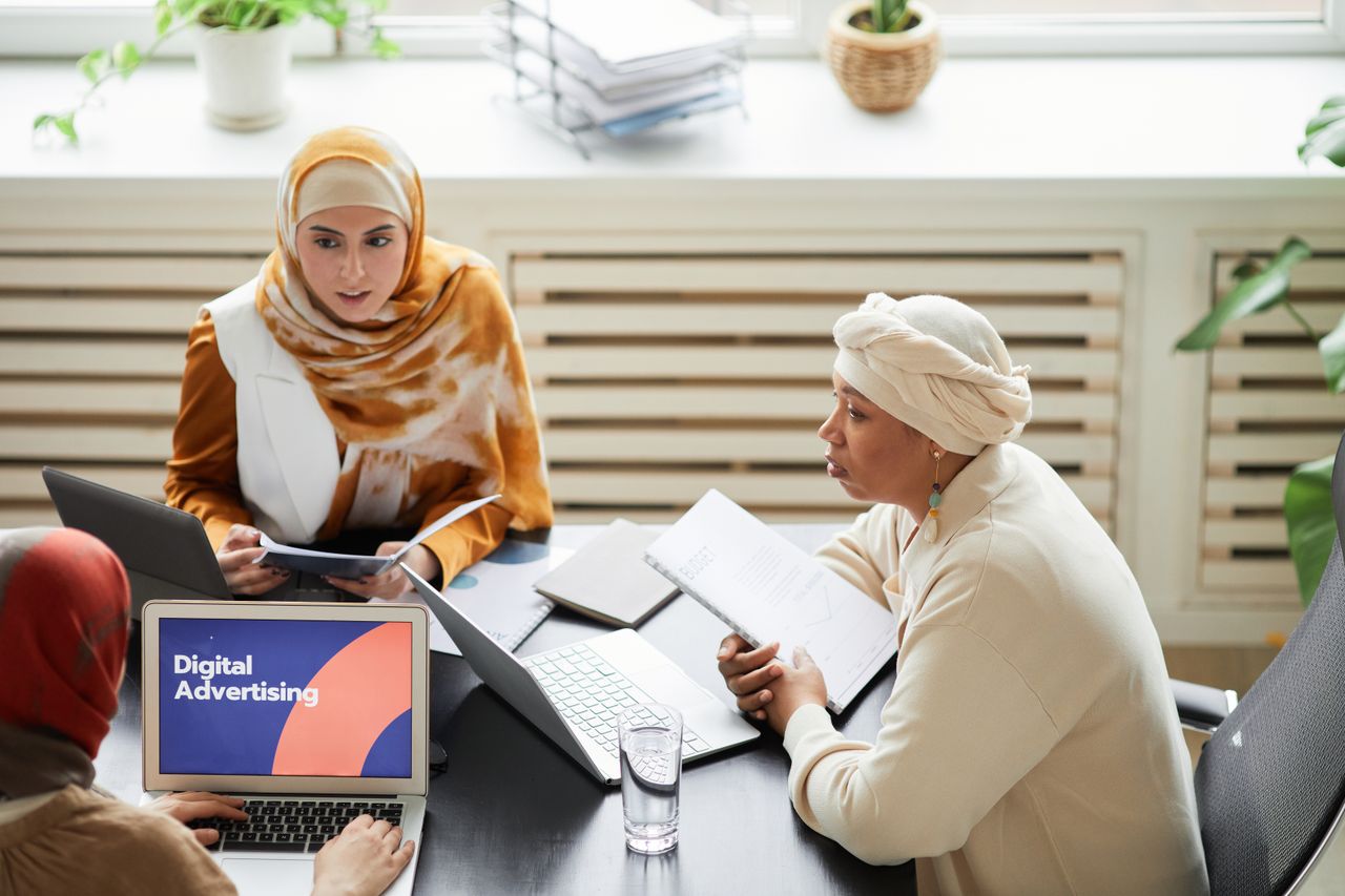 about-01 Three women in hijabs collaborating on digital advertising strategies around a table with laptops.